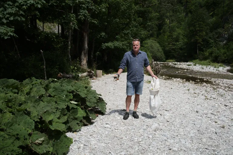 Ein Mann in einem blauen Hemd und Jeansshorts hält eine weiße Tasche in der Hand, während er auf einem Kiesweg in der Nähe eines Flusses steht, mit üppigem Grün und Bäumen im Hintergrund.