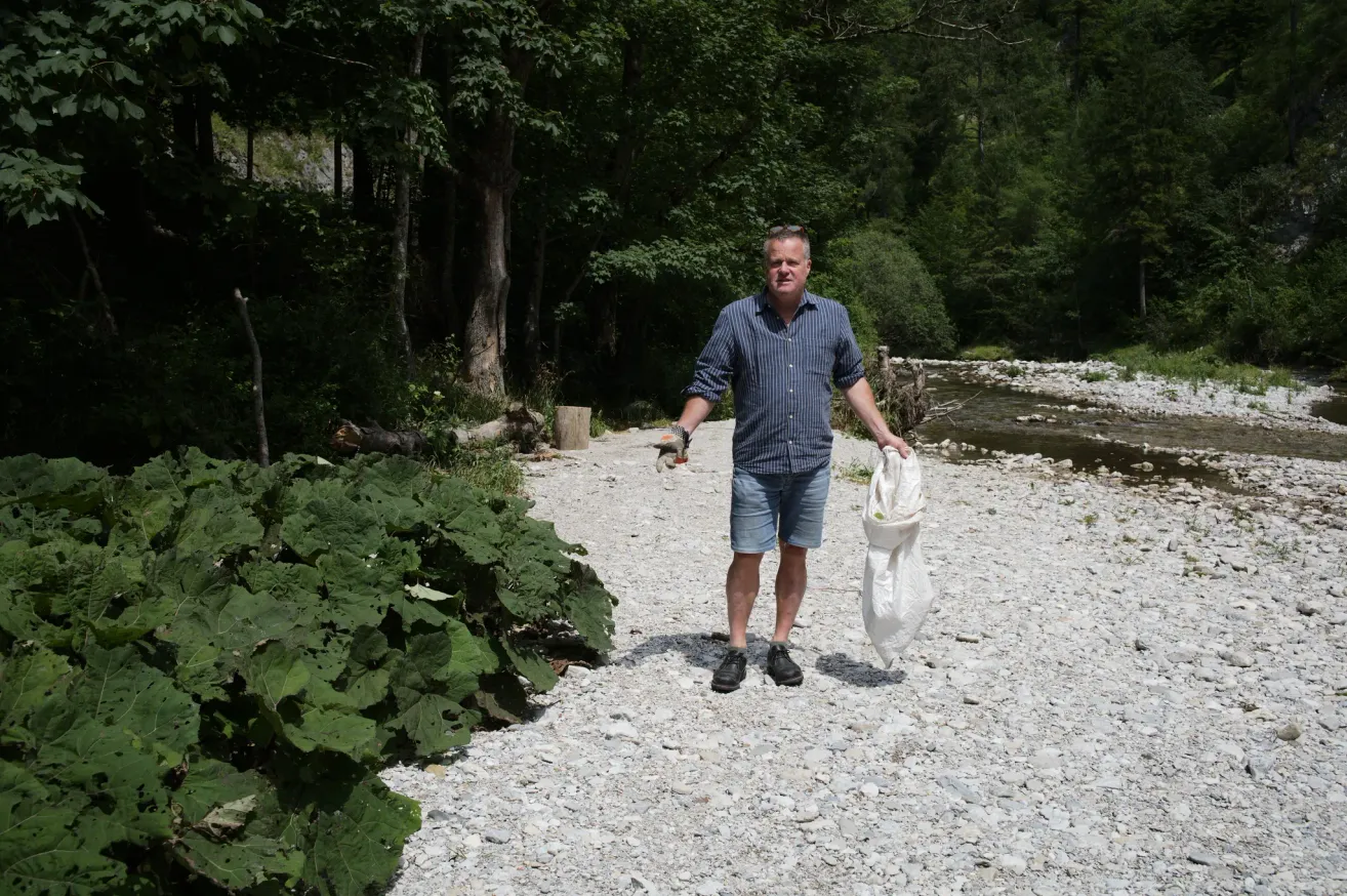 Ein Mann in einem blauen Hemd und Jeansshorts hält eine weiße Tasche in der Hand, während er auf einem Kiesweg in der Nähe eines Flusses steht, mit üppigem Grün und Bäumen im Hintergrund.