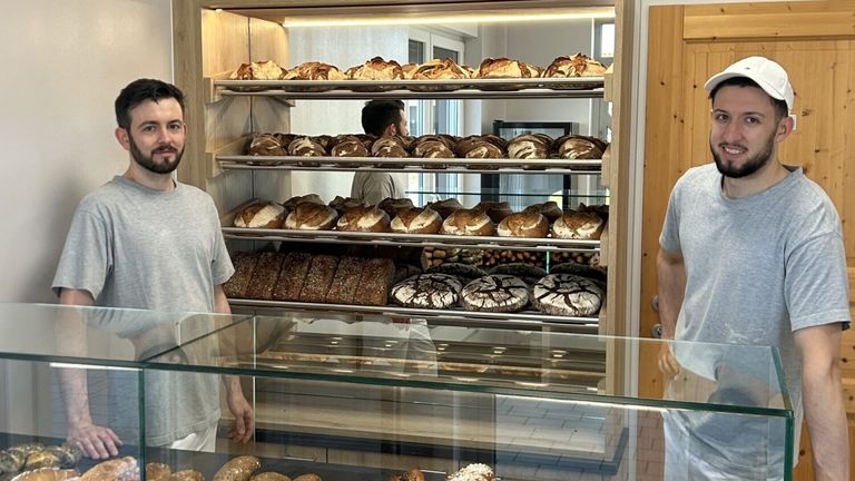 Eine Bäckerei-Vitrine gefüllt mit verschiedenen Brotsorten, einschließlich Laibe und runde Gebäckstücke, unter hellen Lichtern. Ein Mann steht im Hintergrund.