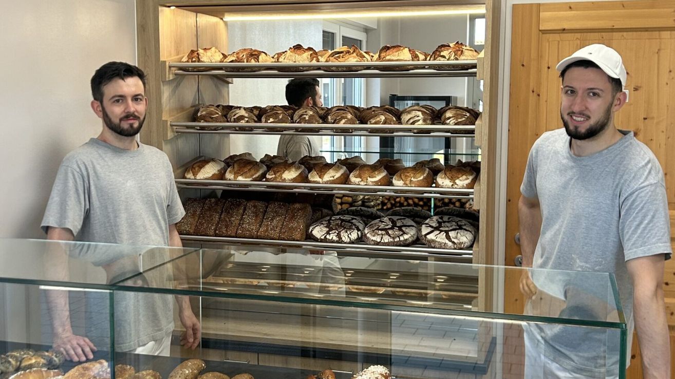 Eine Bäckerei-Vitrine gefüllt mit verschiedenen Brotsorten, einschließlich Laibe und runde Gebäckstücke, unter hellen Lichtern. Ein Mann steht im Hintergrund.