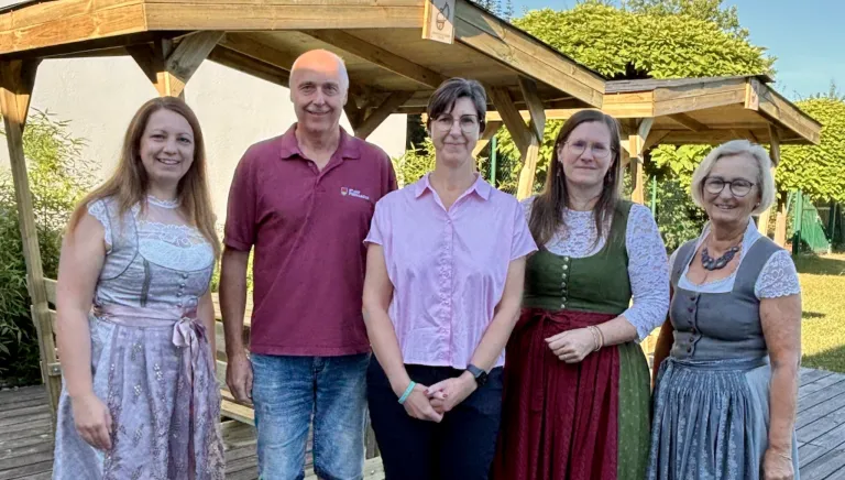 Vier Personen stehen lächelnd vor einer Holzstruktur für ein Foto. Der Mann links trägt ein weinrotes Hemd, und die Frau in der Mitte trägt eine Brille. Die Frau rechts trägt ein traditionelles Kleid.