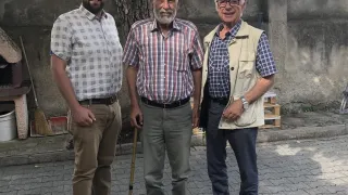Drei Männer stehen auf einem gepflasterten Weg und lächeln. Der Mann auf der linken Seite trägt ein kariertes Hemd und Hose. Der Mann in der Mitte benutzt einen Stock und trägt ein kariertes Hemd. Der Mann auf der rechten Seite trägt eine Weste und eine Brille. Hinter ihnen befindet sich eine Wand mit einem Baumstamm.