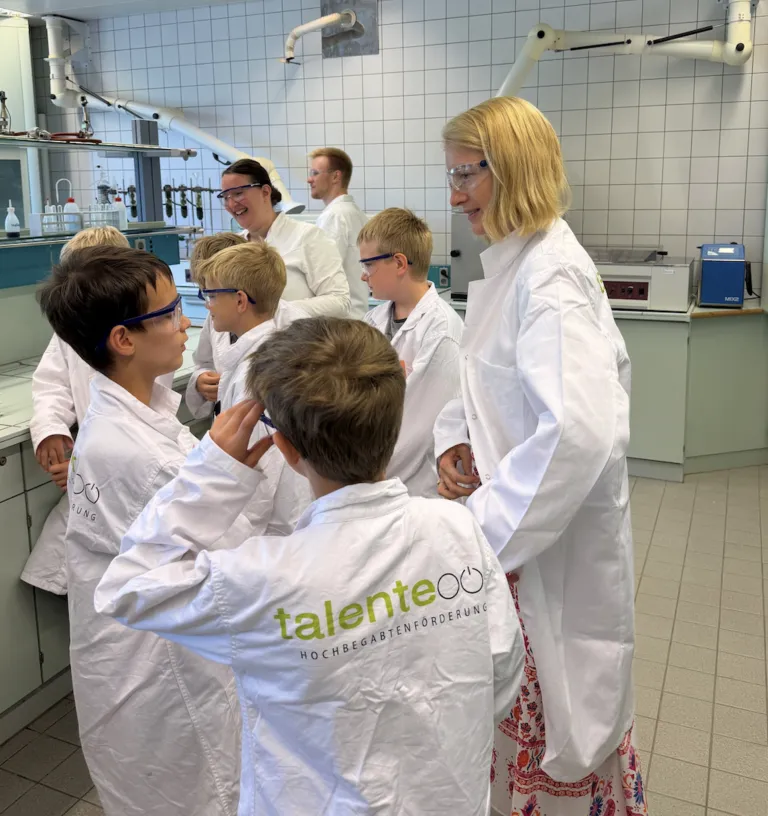 Eine Gruppe von Kindern in Laborkitteln und Schutzbrillen ist um eine Frau in einem Laborkittel versammelt, möglicherweise führen sie ein wissenschaftliches Experiment durch.