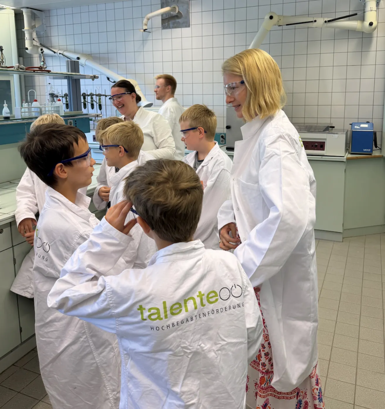 Eine Gruppe von Kindern in Laborkitteln und Schutzbrillen ist um eine Frau in einem Laborkittel versammelt, möglicherweise führen sie ein wissenschaftliches Experiment durch.