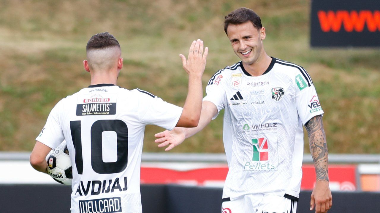 Zwei Fußballspieler feiern ein Tor, einer trägt ein weißes Trikot mit der Nummer 10 und der andere mit einem lächelnden Gesicht und einem Trikot mit verschiedenen Logos.