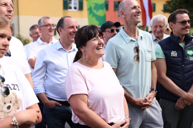 Eine Gruppe von Menschen steht zusammen, lächelt und lacht. Eine Frau in einem rosa T-Shirt steht in der Mitte, mit einem Mann in einem hellblauen Poloshirt auf ihrer rechten Seite und einem anderen Mann in einem weißen Hemd auf ihrer linken Seite. Dahinter stehen weitere Menschen und ein Gebäude mit einem grünen Banner.