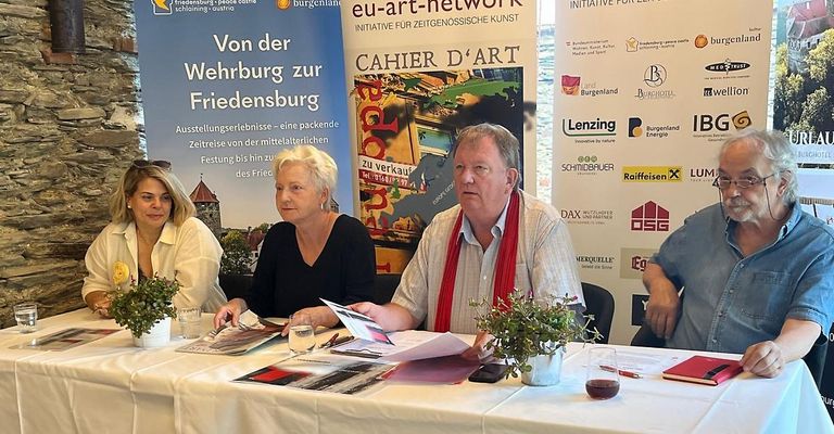 Drei Personen sitzen an einem Tisch mit verschiedenen Papieren und einer Pflanze. Ein Mann liest ein Papier, während eine Frau ihn ansieht. Hinter ihnen ist ein Banner mit dem Text 'CAHIER D'ART'.