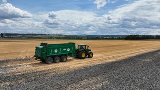 Ein grüner Traktor zieht einen grünen Anhänger durch ein geerntetes Weizenfeld, mit einem bewölkten Himmel und entfernten Windrädern im Hintergrund.