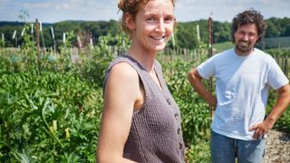 Eine Frau in einem gestrickten Westchen steht in einem Garten mit einem Mann hinter ihr, beide lächeln und schauen zur Kamera.