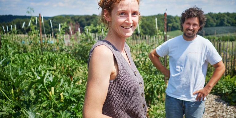 Eine Frau in einem gestrickten Westchen steht in einem Garten mit einem Mann hinter ihr, beide lächeln und schauen zur Kamera.
