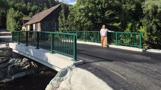 Eine Frau steht auf einer Brücke mit grünen Geländern und blickt auf einen hölzernen Schuppen mit einem Ziegeldach im Hintergrund.