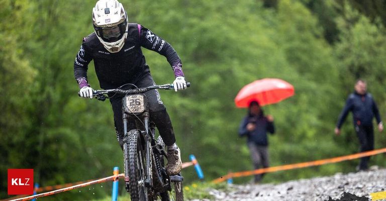 Ein Mountainbiker fährt auf einem schlammigen Trail, trägt einen Helm und Handschuhe. Im Hintergrund hält eine Person einen roten Regenschirm.