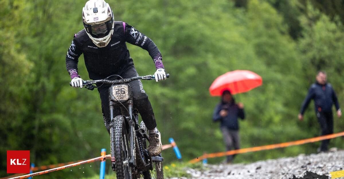 Ein Mountainbiker fährt auf einem schlammigen Trail, trägt einen Helm und Handschuhe. Im Hintergrund hält eine Person einen roten Regenschirm.