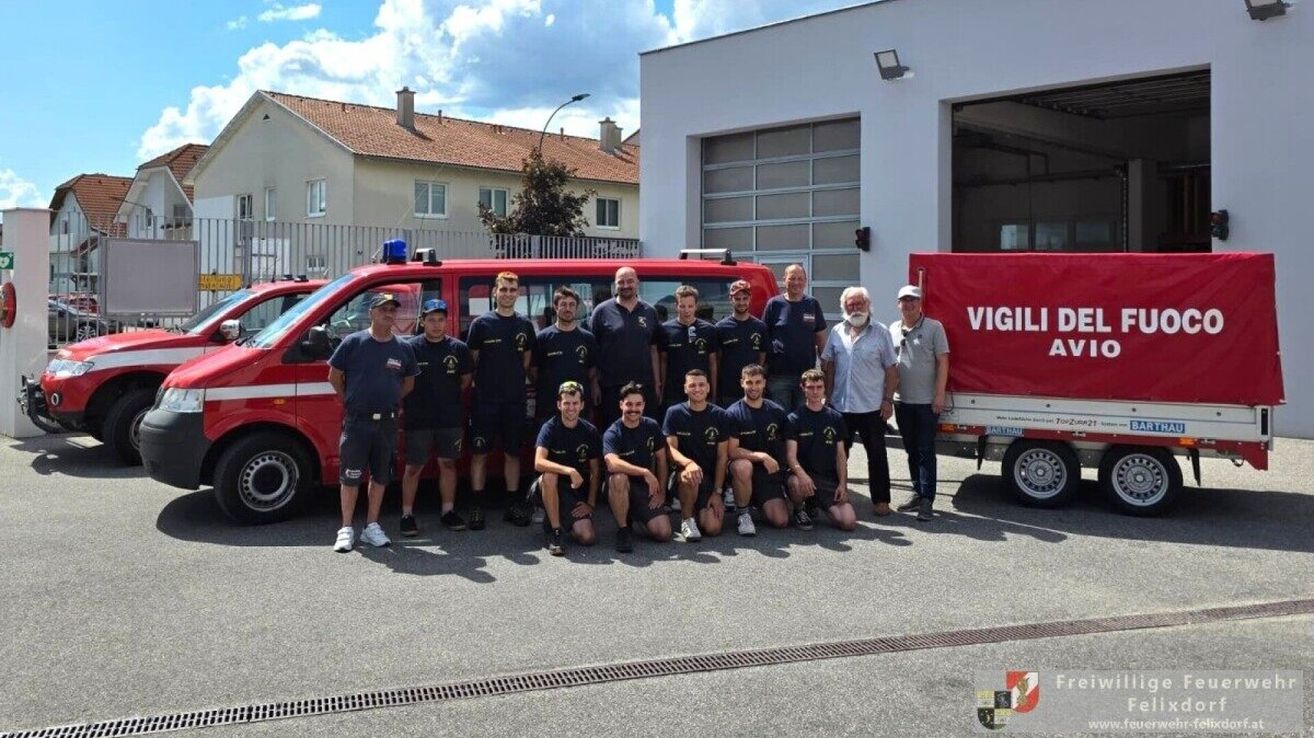 Eine Gruppe Feuerwehrleute in blauen Uniformen und Mützen posiert für ein Foto vor einem roten Feuerwehrauto und einem Gebäude mit einer Garagentür.