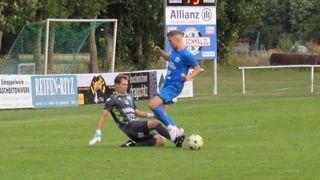 Zwei Fußballspieler sind auf einem Feld, einer in Blau und einer in Schwarz. Der Spieler in Blau versucht, den Ball zu schießen, während der Spieler in Schwarz versucht, ihn zu blockieren. Ein Tor und ein Schild sind im Hintergrund.