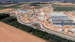Luftaufnahme einer großen Baustelle mit mehreren im Bau befindlichen Gebäuden, Kränen und Fahrzeugen. Die Baustelle ist von Ackerland und Bäumen umgeben.