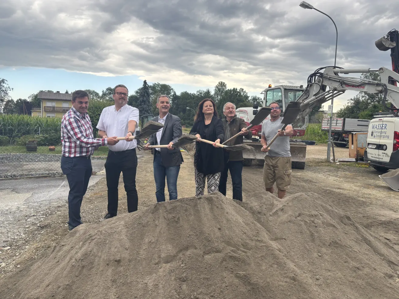 Fünf Personen halten Schaufeln über einem Haufen Erde, wahrscheinlich an einer Baustelle. Dahinter ist ein Baufahrzeug geparkt.
