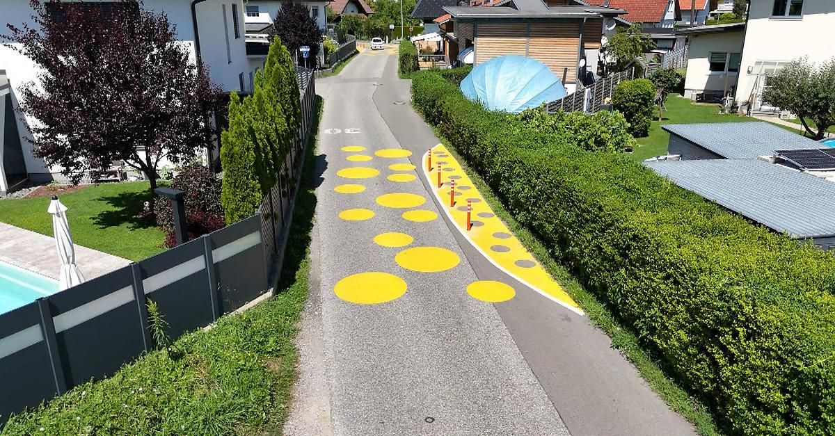 Eine Straße mit gelben Kreisen auf der Fahrbahn, einem Auto in der Ferne und einer blauen Struktur hinter einer Hecke.