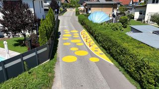Eine Straße mit gelben Kreisen auf der Fahrbahn, einem Auto in der Ferne und einer blauen Struktur hinter einer Hecke.