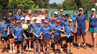 Eine Gruppe von Kindern und Erwachsenen, die blaue Hemden und Shorts tragen, posiert für ein Foto auf einem Tennisplatz mit Tennisschlägern und einem Tennisball.