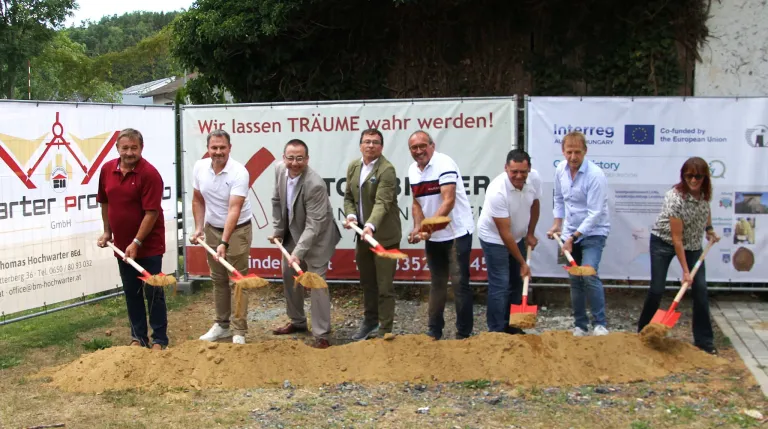 Sechs Männer in weißen Hemden und Hosen stehen in einer Erdgrube mit Schaufeln und lächeln für ein Foto. Hinter ihnen ist ein Banner mit dem Slogan 'Wir lassen Träume wahr werden!'.