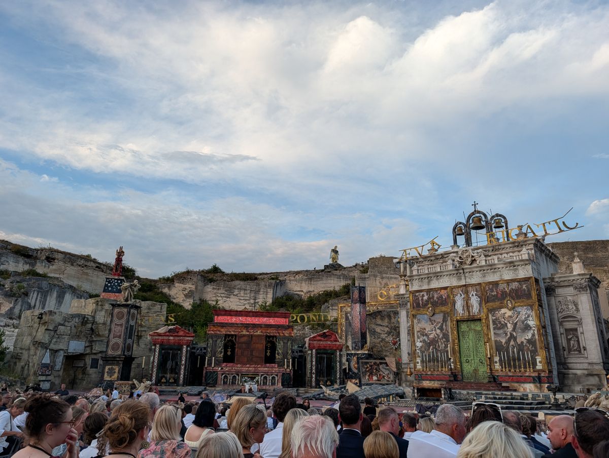 Eine Menschenmenge versammelt sich, um eine Theateraufführung vor dem Hintergrund einer Klippe und einer großen Bühne mit aufwendigen Designs zu sehen.