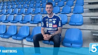 Ein Mann in einem blau-weißen Sporttrikot mit verschiedenen Logos sitzt auf einer blauen Tribüne in einem leeren Stadion.