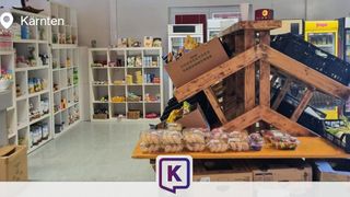 Ein gut bestückter Laden mit Regalen voller verschiedener Lebensmittel, einem hölzernen Displayständer mit verpackten Snacks und einem Kühlschrank im Hintergrund.