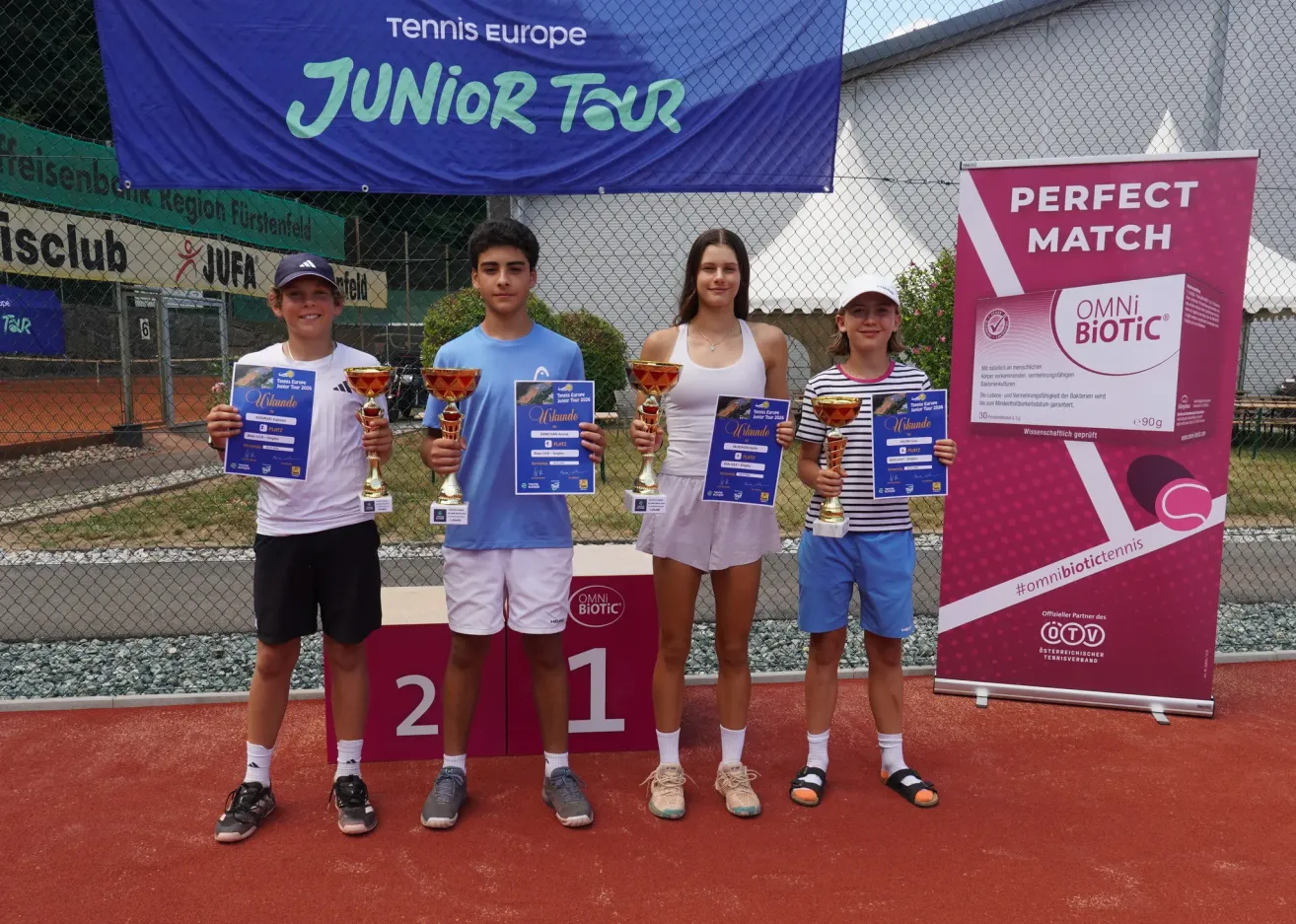 Vier junge Tennisspieler stehen auf einem roten Platz und halten Pokale. Sie lächeln und scheinen für ein Foto zu posieren. Hinter ihnen ist ein Zaun mit einem Banner, auf dem 'Junior Tour' steht.
