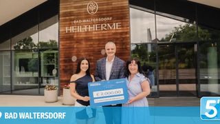 Drei Personen stehen vor einem Gebäude mit dem Schild Bad Waltersdorf Heiltherme. Der Mann und die beiden Frauen lächeln und halten einen großen blauen Scheck über 2.000 Euro.