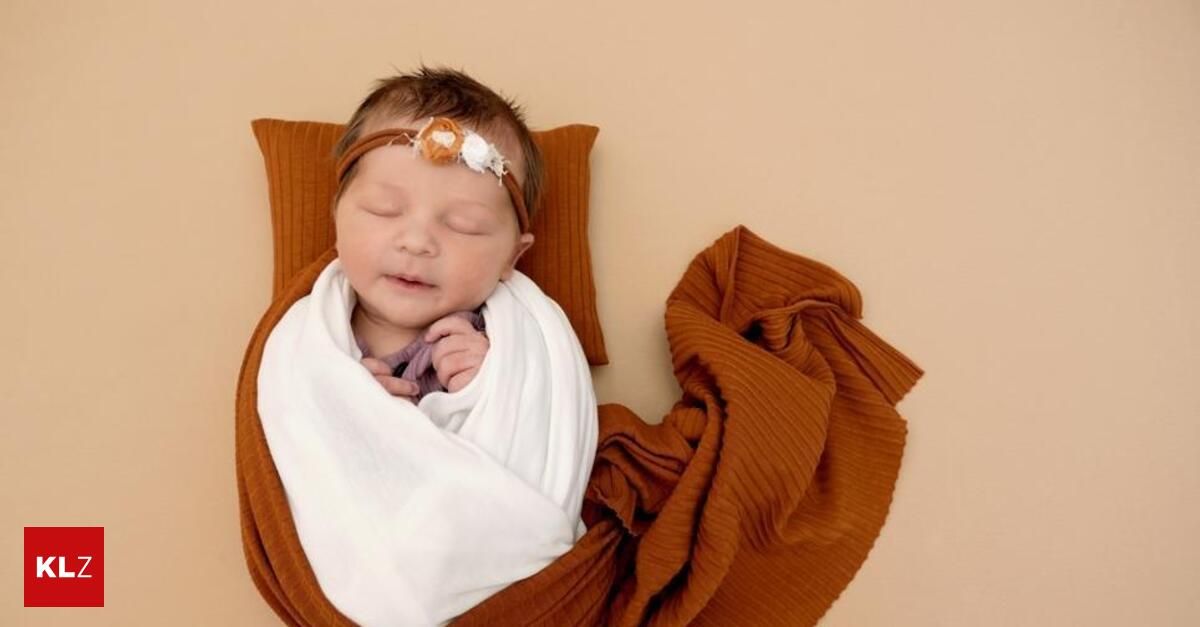 Ein neugeborenes Babymädchen schläft friedlich, eingewickelt in einen weißen Stoff und einen braunen Decken, mit einem Stirnband auf dem Kopf.
