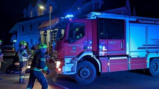 Ein Feuerwehrmann in voller Montur geht an einem roten Feuerwehrwagen mit der Nummer HLF3 auf der Seite vorbei, der nachts vor einem Gebäude geparkt ist.