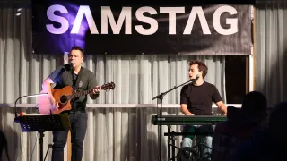 Zwei Männer treten auf der Bühne auf, einer spielt Gitarre und der andere spielt Keyboard. Ein Banner mit der Aufschrift 'SAMSTAG' ist über ihnen zu sehen.