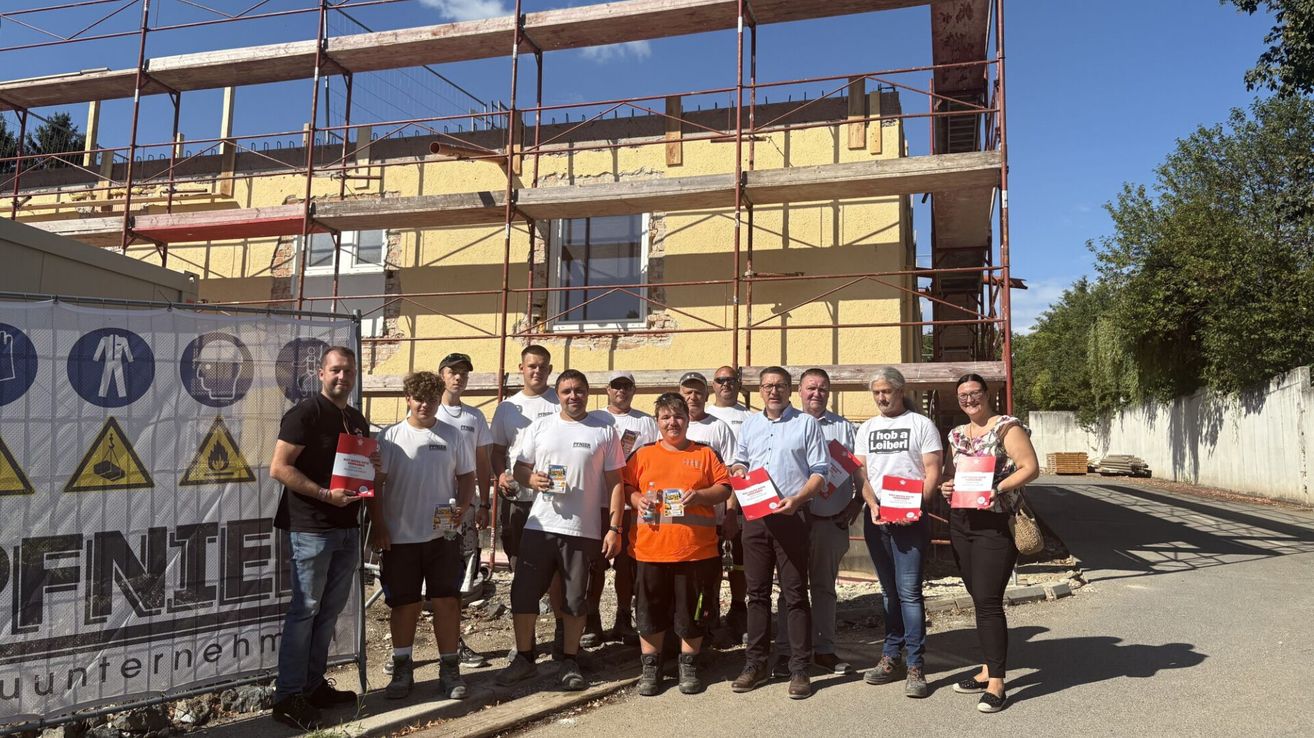 Eine Gruppe von Menschen vor einem im Bau befindlichen Gebäude, die rote Zertifikate halten und für ein Foto posieren.