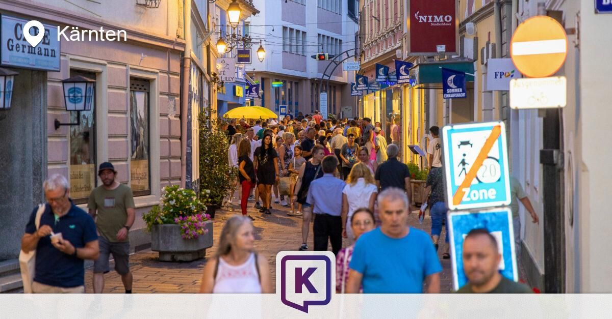 Eine belebte Straße mit Fußgängern und Geschäften auf beiden Seiten, eines mit einem Schild 'NIMO' und ein anderes mit einer blauen Flagge.
