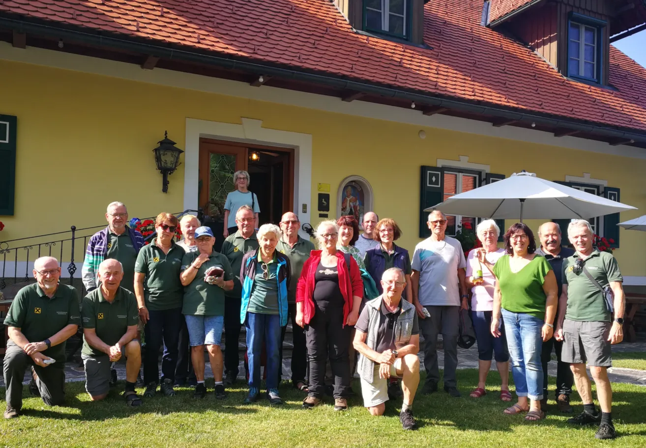 Eine Gruppe von Menschen, meist älterer Menschen, posiert für ein Foto vor einem gelben Haus mit einem roten Dach. Einige tragen Sonnenbrillen und Hüte.