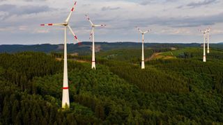 Drei Windturbinen stehen in einem üppigen grünen Wald unter einem bewölkten Himmel. Die Turbinen haben weiße Blätter mit roten Streifen.