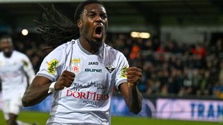 Ein Fußballspieler feiert ein Tor mit offenem Mund und erhobenen Fäusten vor einem jubelnden Publikum.