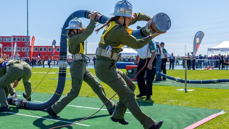 Zwei Frauen in grünen Uniformen und Helmen ziehen an großen Schläuchen, möglicherweise in einem Wettbewerb. Sie befinden sich auf einem grünen Feld mit einer Menge im Hintergrund.