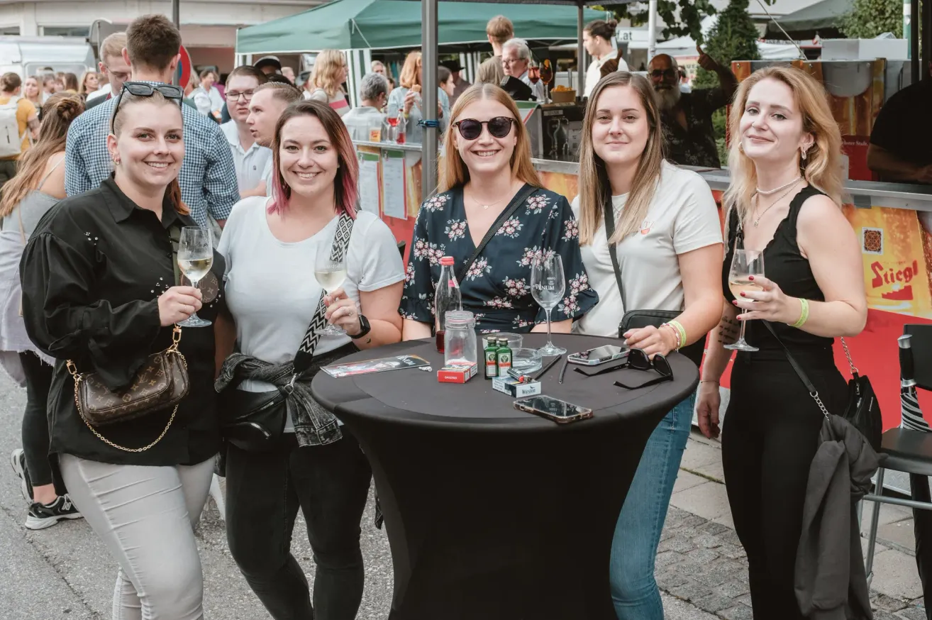 Fünf Frauen stehen um einen Tisch bei einer Veranstaltung im Freien, lächeln und halten Weingläser. Dahinter ist ein Imbissstand mit einem grünen Zelt zu sehen, und mehrere Leute sind unterwegs.