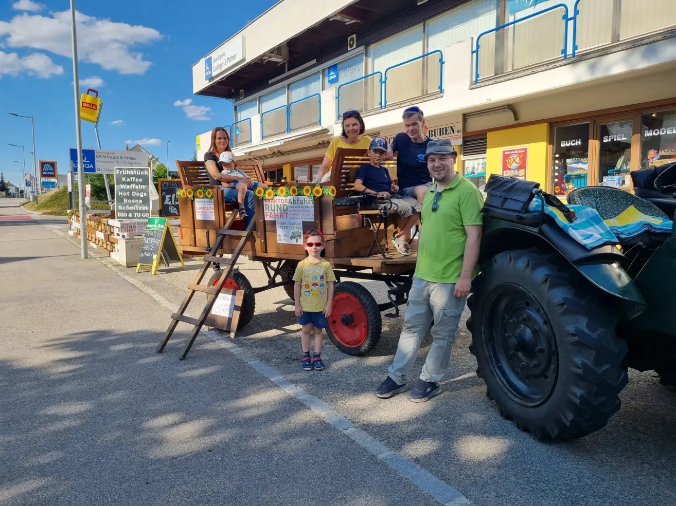Eine Familie mit sechs Personen, darunter zwei Kinder, sitzt auf einem von einem Traktor gezogenen Holzwagen. Sie posieren für ein Foto vor einem Gebäude.
