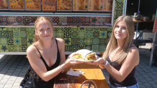 Zwei Frauen sitzen an einem Tisch draußen, lächeln und halten einen Teller mit Essen. Hinter ihnen steht ein Imbisswagen mit bunten Kacheln und Schildern für verschiedene mexikanische Gerichte.