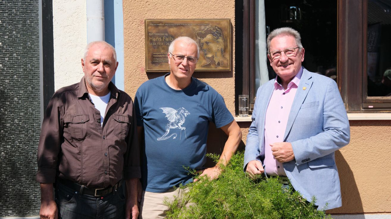 Drei Männer stehen vor einem Gebäude mit einer Plakette dahinter. Der Mann links trägt ein braunes Hemd, der Mann in der Mitte ein blaues Hemd mit Drachendesign und der Mann rechts einen blauen Anzug.