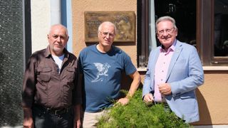 Drei Männer stehen vor einem Gebäude mit einer Plakette dahinter. Der Mann links trägt ein braunes Hemd, der Mann in der Mitte ein blaues Hemd mit Drachendesign und der Mann rechts einen blauen Anzug.