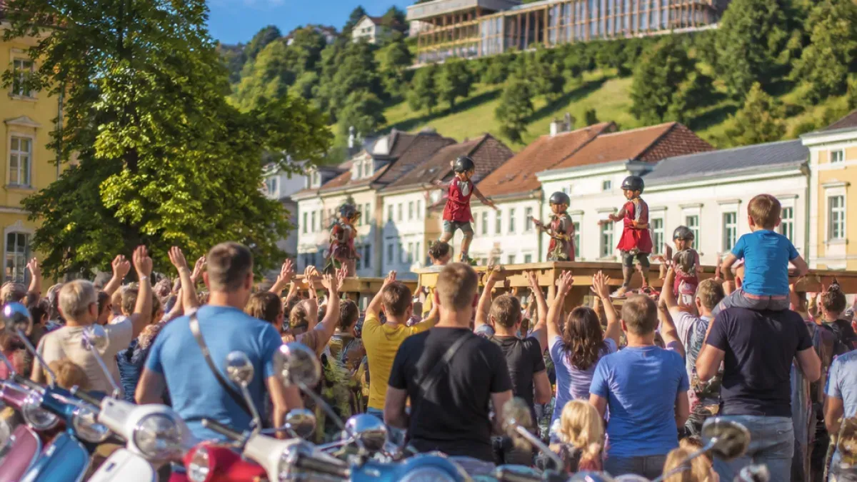 A group of children wearing helmets perform on a stage in front of a crowd. The children are standing on a wooden platform with a building in the background.