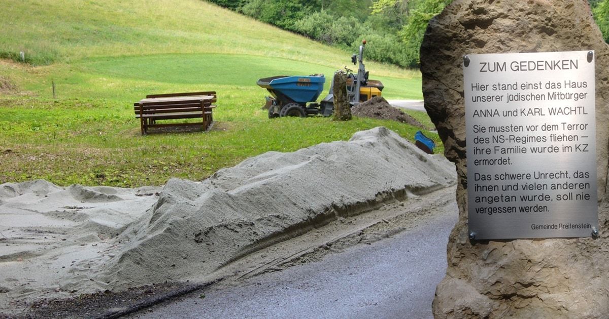 A construction site with a pile of sand and a machinery vehicle in a grassy area. A wooden bench is nearby. A signboard with text is on the right side.