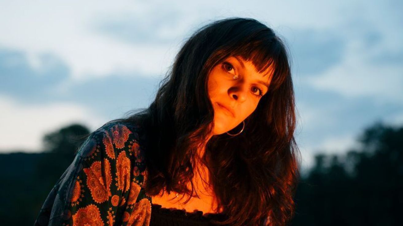 A woman with long black hair and bangs, wearing a patterned blouse, poses for a photo with a sunset in the background.