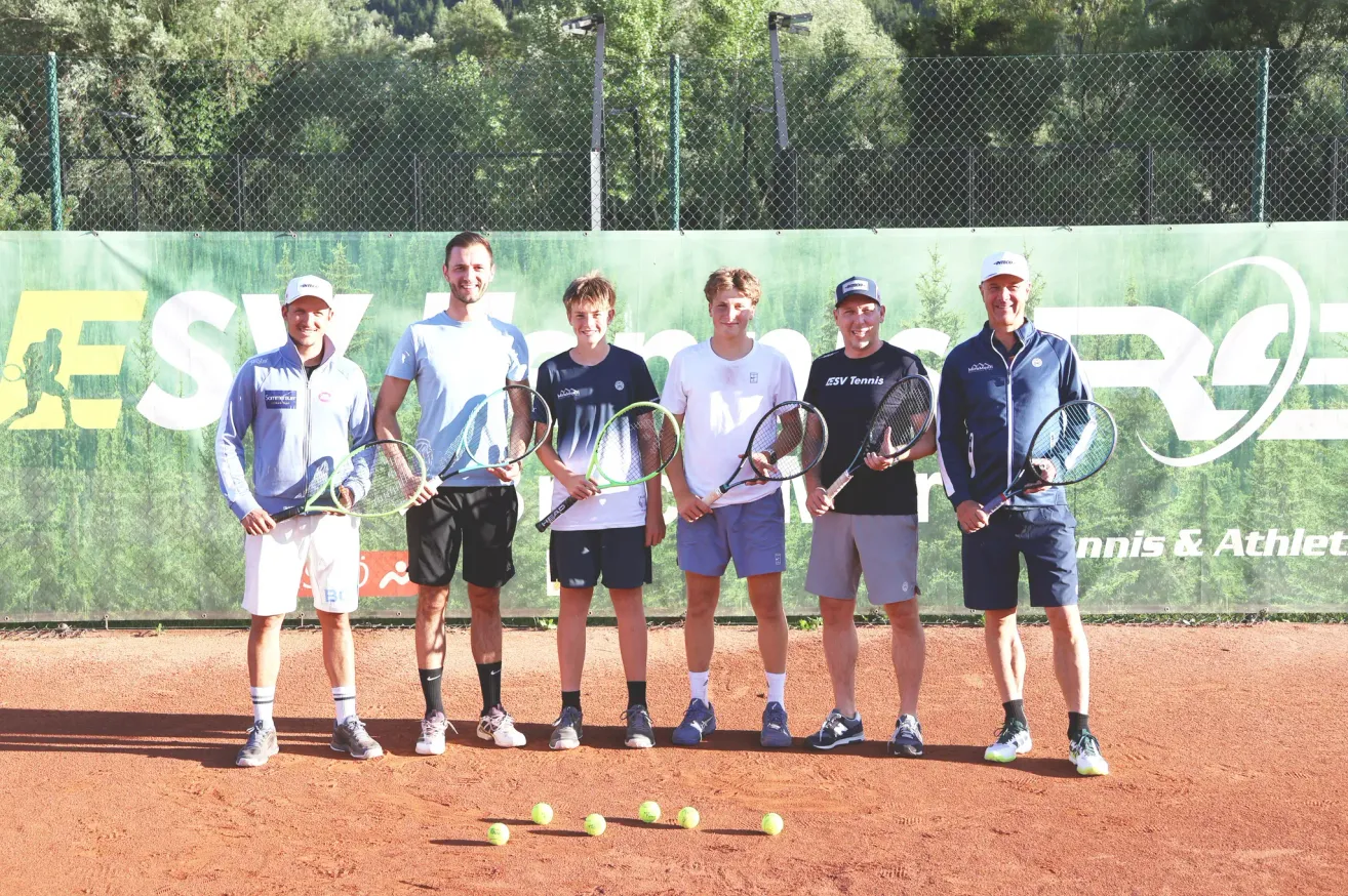 Five men stand on a tennis court with tennis rackets in hand, smiling for a photo. Behind them is a green banner with white letters.