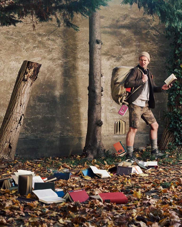 A man wearing a beanie and a backpack is standing in a park with fallen leaves and books around him, holding a book in his hand.