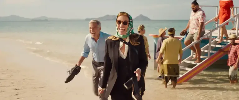 A woman in a green headscarf and sunglasses walks on a beach with a man and other people in the background.